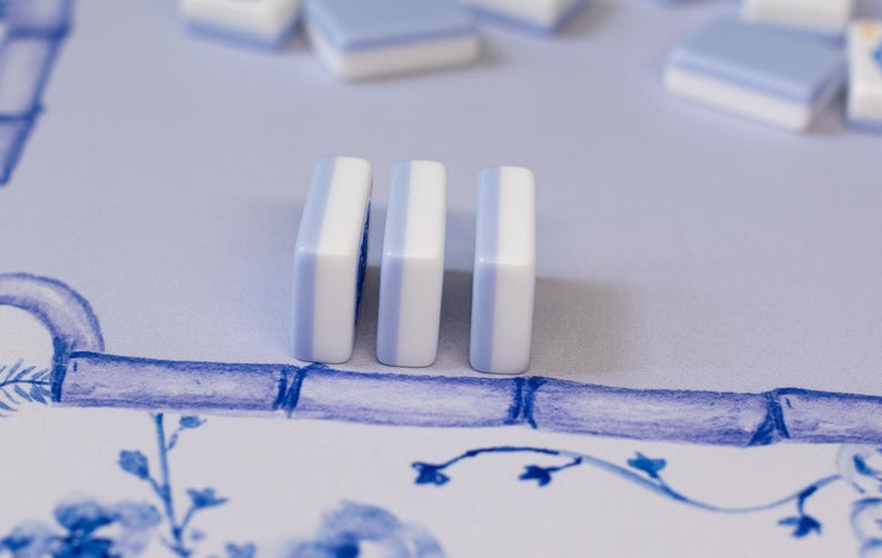 Puede incluir: Tres fichas de juego rectangulares, blancas y azul claro, se alzan sobre una superficie estampada. Hay fichas adicionales dispersas en el fondo. El dise&ntilde;o presenta elementos florales y de estilo bamb&uacute; azules, creando una est&eacute;tica cohesiva.