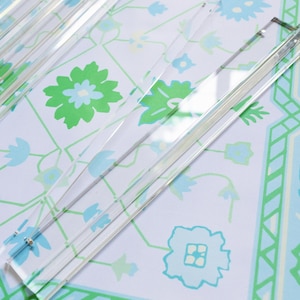 May include: Clear acrylic place card holders arranged on a patterned surface. The holders are rectangular with a slot for cards. The background features a white, blue, and green floral design, creating a fresh, decorative aesthetic.
