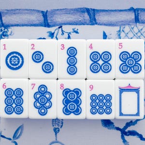 Puede incluir: Un juego de fichas de Mahjong blancas con detalles azules y rosas. Las fichas est&aacute;n numeradas del 1 al 9, con varios dise&ntilde;os circulares azules. El fondo presenta un patr&oacute;n floral azul y blanco.