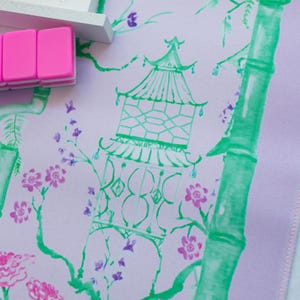 K&ouml;nnte beinhalten: Ein helllila Stoff mit einem gr&uuml;nen Bambus-Design und rosa und violetten Blumen. Eine gr&uuml;ne Pagode mit wei&szlig;em Umriss befindet sich in der Mitte des Stoffes.