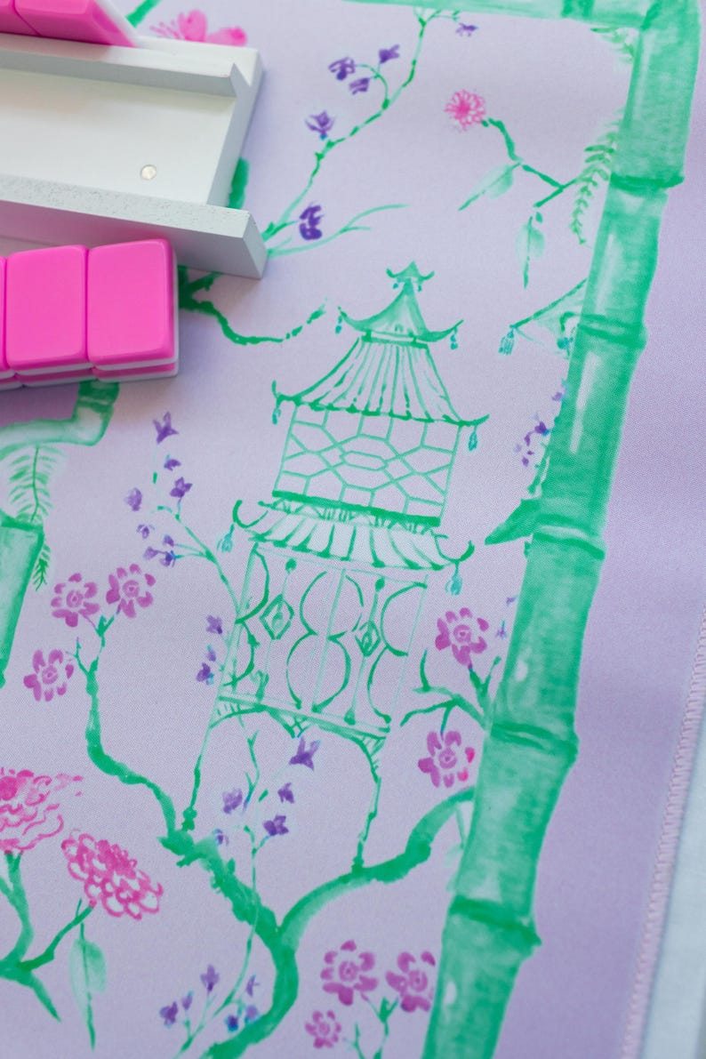 K&ouml;nnte beinhalten: Ein helllila Stoff mit einem gr&uuml;nen Bambus-Design und rosa und violetten Blumen. Eine gr&uuml;ne Pagode mit wei&szlig;em Umriss befindet sich in der Mitte des Stoffes.