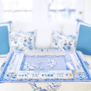 Puede incluir: Un juego de Mahjong azul y blanco sobre una mesa con un mantel floral. El juego incluye fichas y soportes, con una superficie de juego azul claro. Cojines decorativos en el fondo realzan la est&eacute;tica. El tema general es un juego cl&aacute;sico con un toque de elegancia.