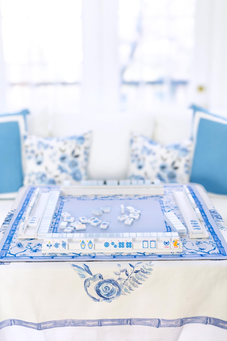 Puede incluir: Un juego de Mahjong azul y blanco sobre una mesa con un mantel floral. El juego incluye fichas y soportes, con una superficie de juego azul claro. Cojines decorativos en el fondo realzan la est&eacute;tica. El tema general es un juego cl&aacute;sico con un toque de elegancia.