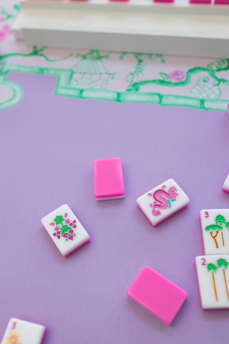 K&ouml;nnte beinhalten: Ein Satz rosa und wei&szlig;er Mahjong-Steine mit verschiedenen Symbolen und Zahlen. Die Steine liegen auf einer violetten Oberfl&auml;che.
