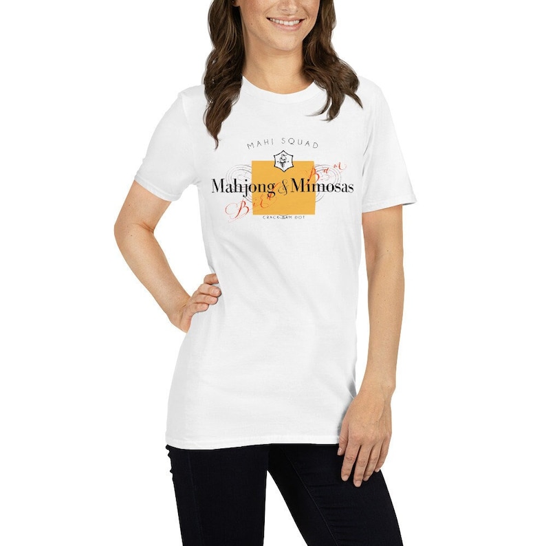May include: White t-shirt with a graphic design of a champagne bottle label that says "Mahjong & Mimosas Brunch Squad".