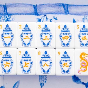 Puede incluir: Diez fichas de Mahjong blancas con dise&ntilde;os de jarrones florales azules y blancos y n&uacute;meros chinos naranjas. Una ficha presenta un drag&oacute;n naranja. Las fichas est&aacute;n dispuestas sobre una superficie con estampado floral azul y blanco.