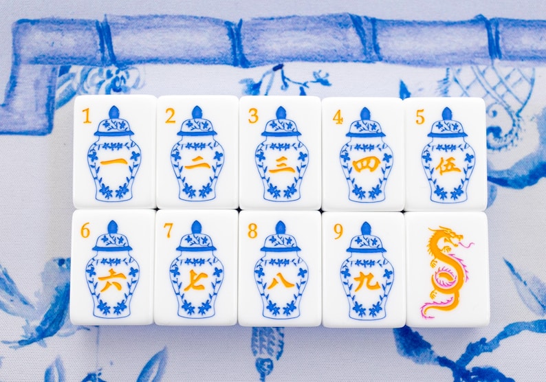 Puede incluir: Diez fichas de Mahjong blancas con dise&ntilde;os de jarrones florales azules y blancos y n&uacute;meros chinos naranjas. Una ficha presenta un drag&oacute;n naranja. Las fichas est&aacute;n dispuestas sobre una superficie con estampado floral azul y blanco.