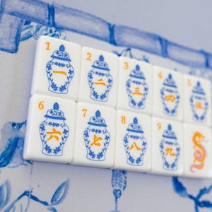 Puede incluir: Un conjunto de fichas de Mahjong blancas con dise&ntilde;os azules y naranjas. Cada ficha presenta una ilustraci&oacute;n de un jarr&oacute;n azul y un n&uacute;mero o s&iacute;mbolo en naranja. Las fichas est&aacute;n dispuestas sobre un fondo floral azul y blanco.