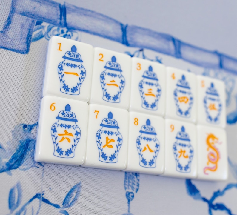 Puede incluir: Un conjunto de fichas de Mahjong blancas con dise&ntilde;os azules y naranjas. Cada ficha presenta una ilustraci&oacute;n de un jarr&oacute;n azul y un n&uacute;mero o s&iacute;mbolo en naranja. Las fichas est&aacute;n dispuestas sobre un fondo floral azul y blanco.