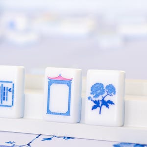 Puede incluir: Primer plano de tres fichas de Mahjong blancas con dise&ntilde;os azules y rosas. Una ficha presenta un dise&ntilde;o floral, otra una pagoda y la tercera un patr&oacute;n geom&eacute;trico. Las fichas se exhiben sobre una superficie blanca, lo que sugiere un juego o un objeto decorativo.