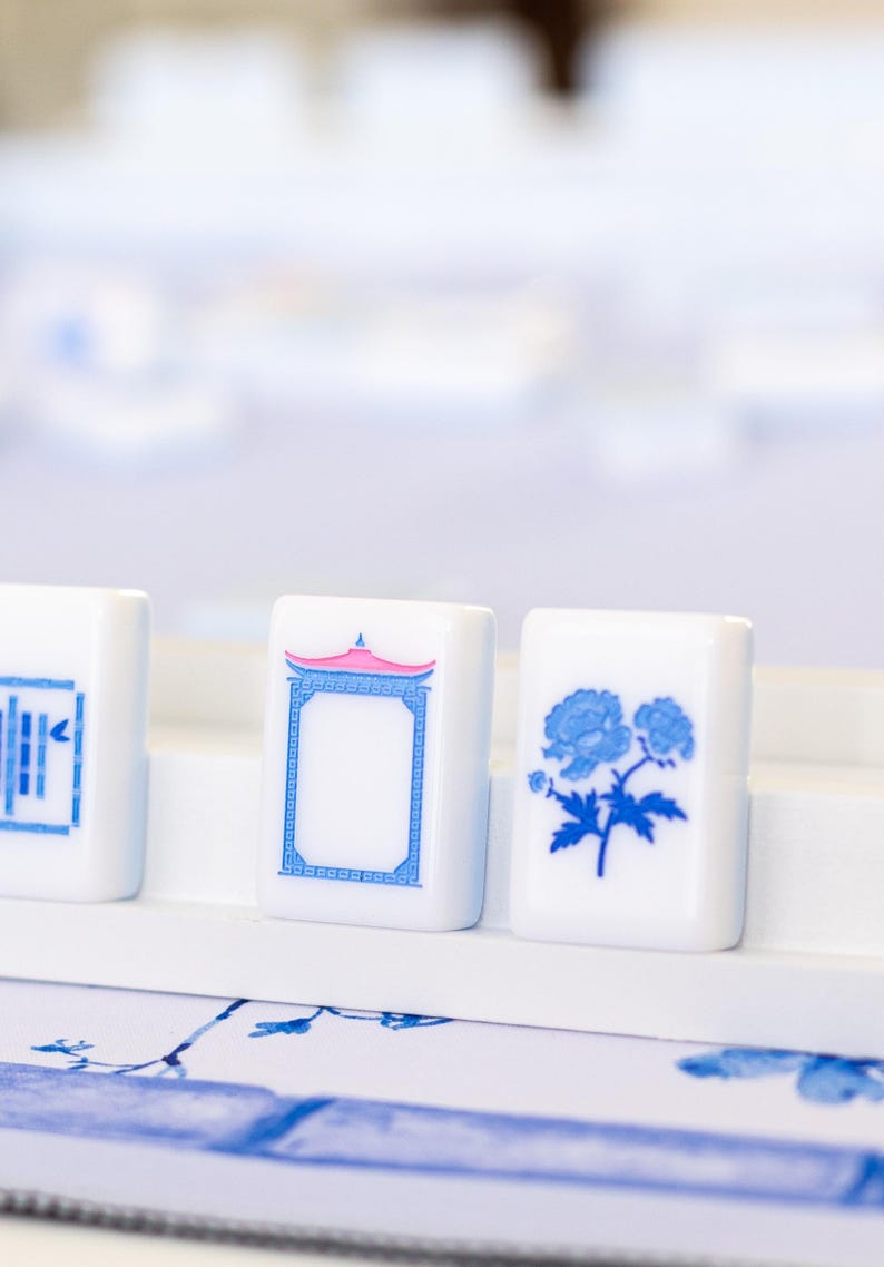 Puede incluir: Primer plano de tres fichas de Mahjong blancas con dise&ntilde;os azules y rosas. Una ficha presenta un dise&ntilde;o floral, otra una pagoda y la tercera un patr&oacute;n geom&eacute;trico. Las fichas se exhiben sobre una superficie blanca, lo que sugiere un juego o un objeto decorativo.