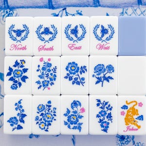 Puede incluir: Un conjunto de fichas de Mahjong blancas con dise&ntilde;os florales y de abejas azules. Las fichas est&aacute;n dispuestas sobre una superficie con estampado floral azul y blanco. Algunas fichas tienen las palabras "Norte", "Sur", "Este" y "Oeste".