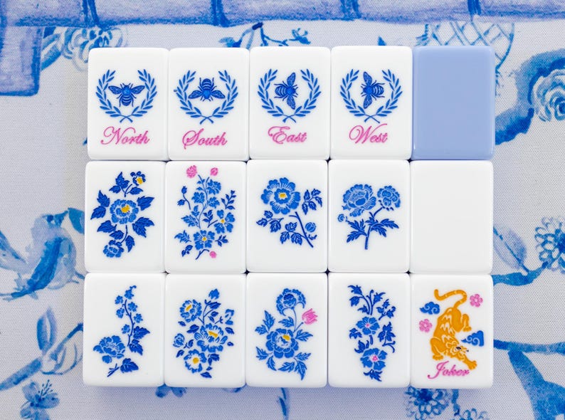 Puede incluir: Un conjunto de fichas de Mahjong blancas con dise&ntilde;os florales y de abejas azules. Las fichas est&aacute;n dispuestas sobre una superficie con estampado floral azul y blanco. Algunas fichas tienen las palabras "Norte", "Sur", "Este" y "Oeste".
