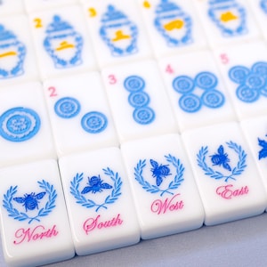 Puede incluir: Primer plano de un juego de fichas de Mahjong blancas. Las fichas presentan dise&ntilde;os azules y rosas, incluidos patrones florales, n&uacute;meros e indicadores direccionales como "Norte", "Sur", "Este" y "Oeste".