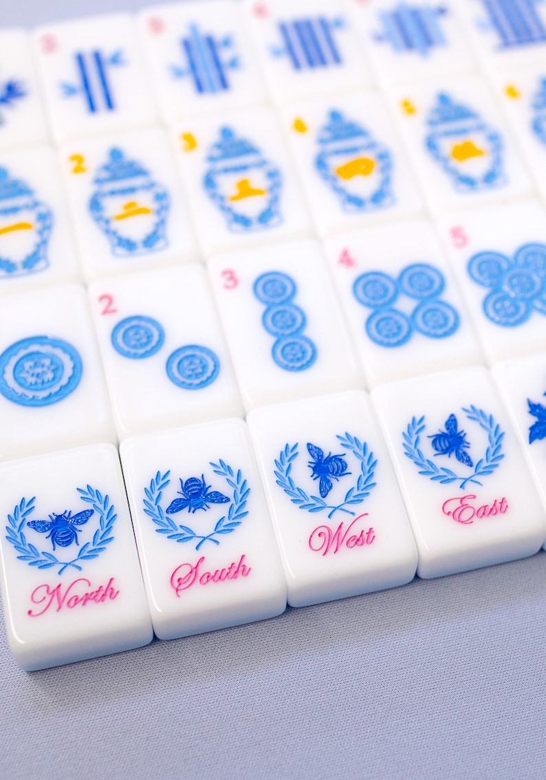Puede incluir: Primer plano de un juego de fichas de Mahjong blancas. Las fichas presentan dise&ntilde;os azules y rosas, incluidos patrones florales, n&uacute;meros e indicadores direccionales como "Norte", "Sur", "Este" y "Oeste".
