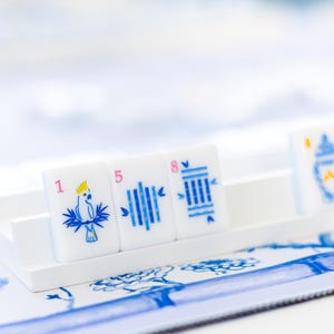 Puede incluir: Azulejos blancos con ilustraciones y n&uacute;meros azules, dispuestos sobre una bandeja blanca. Los azulejos presentan un loro, l&iacute;neas verticales y una pila de l&iacute;neas. El fondo incluye un estampado floral azul y blanco.