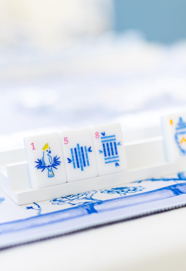 Puede incluir: Azulejos blancos con ilustraciones y n&uacute;meros azules, dispuestos sobre una bandeja blanca. Los azulejos presentan un loro, l&iacute;neas verticales y una pila de l&iacute;neas. El fondo incluye un estampado floral azul y blanco.