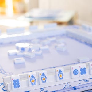 Puede incluir: Un juego de Mahjong con fichas blancas con dise&ntilde;os azules y naranjas. Las fichas est&aacute;n dispuestas sobre una bandeja blanca, con un fondo con estampado floral azul y blanco. El juego est&aacute; configurado y listo para jugar.