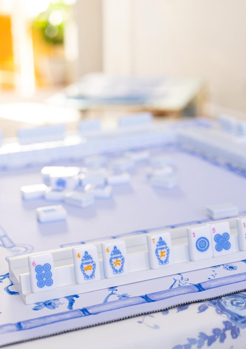 Puede incluir: Un juego de Mahjong con fichas blancas con dise&ntilde;os azules y naranjas. Las fichas est&aacute;n dispuestas sobre una bandeja blanca, con un fondo con estampado floral azul y blanco. El juego est&aacute; configurado y listo para jugar.