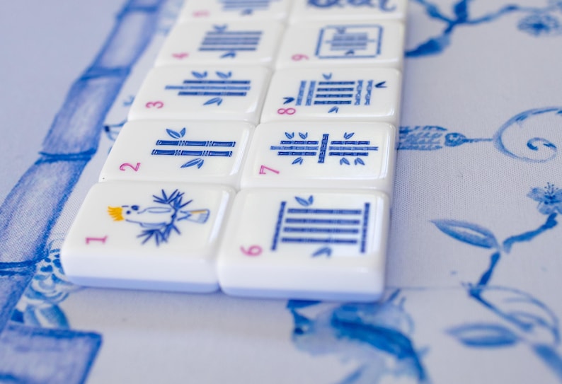 Puede incluir: Primer plano de fichas de Mahjong blancas con ilustraciones y n&uacute;meros azules. Las fichas presentan dise&ntilde;os de bamb&uacute;, p&aacute;jaros y geom&eacute;tricos. Las fichas est&aacute;n dispuestas sobre una superficie con estampado floral azul y blanco.