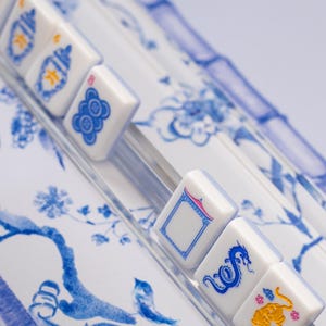 Puede incluir: Primer plano de fichas de Mahjong blancas con s&iacute;mbolos y caracteres azules, incluyendo un drag&oacute;n y un tigre. Las fichas est&aacute;n dispuestas sobre una superficie transparente e inclinada, sobre un fondo de patrones florales azules y blancos.