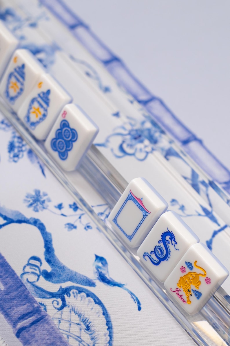 Puede incluir: Primer plano de fichas de Mahjong blancas con s&iacute;mbolos y caracteres azules, incluyendo un drag&oacute;n y un tigre. Las fichas est&aacute;n dispuestas sobre una superficie transparente e inclinada, sobre un fondo de patrones florales azules y blancos.