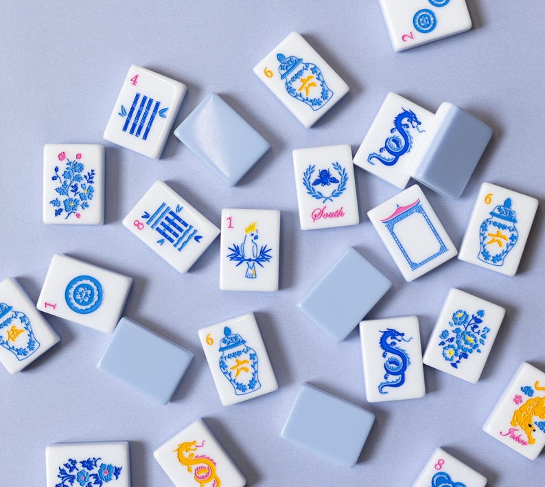 Puede incluir: Vista a&eacute;rea de una colecci&oacute;n de fichas de Mahjong blancas y azules dispersas sobre una superficie azul claro. Las fichas presentan varias ilustraciones azules, incluyendo patrones florales, dragones y dise&ntilde;os geom&eacute;tricos. Algunas fichas tienen n&uacute;meros.