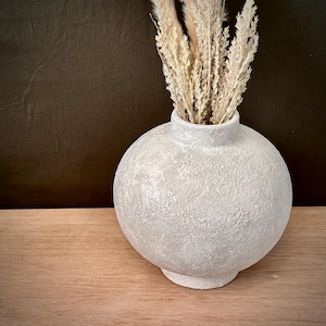 May include: A textured, round, off-white vase with a narrow neck, holding dried beige ornamental grass. The vase sits on a wooden surface against a dark brown backdrop. The vase has a rough, stucco-like finish.