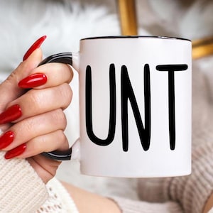 Può includere: Una tazza in ceramica bianca con un bordo nero e la parola "UNT" stampata in nero.