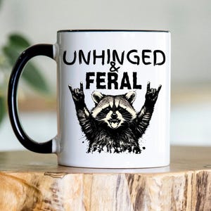 Puede incluir: Taza de cer&aacute;mica blanca con asa y borde negros. La taza presenta una ilustraci&oacute;n de mapache con el texto "UNHINGED & FERAL" en negro. El mapache hace un gesto de rock and roll con la mano.