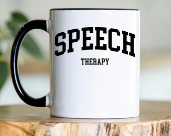 Può includere: Tazza in ceramica bianca con manico e bordo neri. La tazza presenta le parole "SPEECH THERAPY" in grassetto nero, in stile collegiale. La tazza è esposta su una superficie di legno.