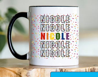 Może przedstawiać: Biały ceramiczny kubek do kawy z czarnym uchwytem. Kubek ma napisane imię "Nicole" pięć razy w kolorach tęczy na białym tle z kolorowymi posypkami. Tekst "PREZENT DO SPERSONALIZOWANIA" jest napisany niebieskim kolorem na białym tle.
