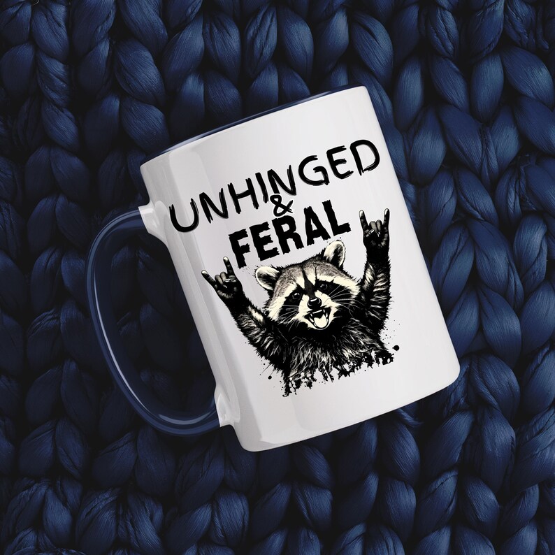 Puede incluir: Taza de cer&aacute;mica blanca con asa azul oscuro. La taza tiene el texto "UNHINGED & FERAL" sobre una ilustraci&oacute;n en blanco y negro de un mapache haciendo un gesto de rock and roll. Fondo de punto azul oscuro.