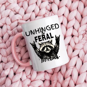 Puede incluir: Taza de cer&aacute;mica blanca con asa e interior rosa. La taza presenta una ilustraci&oacute;n en blanco y negro de un mapache con los brazos levantados en un gesto de rock and roll. El texto "UNHINGED & FERAL" est&aacute; impreso encima del mapache.
