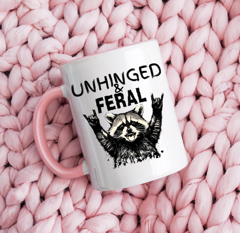 Puede incluir: Taza de cer&aacute;mica blanca con asa e interior rosa. La taza presenta una ilustraci&oacute;n en blanco y negro de un mapache con los brazos levantados en un gesto de rock and roll. El texto "UNHINGED & FERAL" est&aacute; impreso encima del mapache.