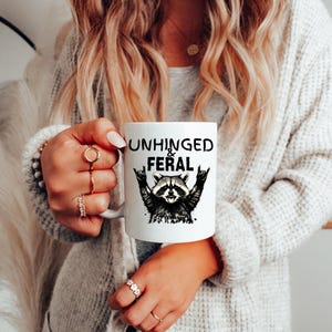 Puede incluir: Taza de cer&aacute;mica blanca con las palabras "UNHINGED & FERAL" en negro, junto con una ilustraci&oacute;n en blanco y negro de un mapache. La taza tiene un asa blanca.
