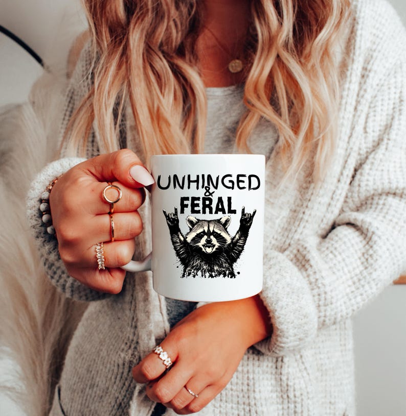 Puede incluir: Taza de cer&aacute;mica blanca con las palabras "UNHINGED & FERAL" en negro, junto con una ilustraci&oacute;n en blanco y negro de un mapache. La taza tiene un asa blanca.