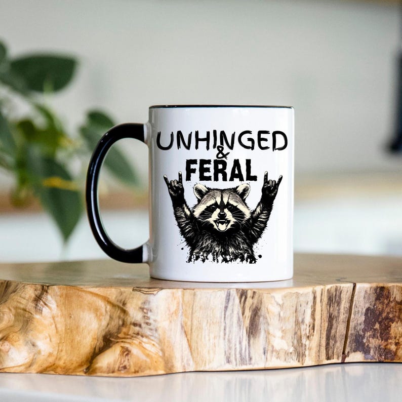 Puede incluir: Taza de cer&aacute;mica blanca con asa y borde negros. La taza presenta una ilustraci&oacute;n de mapache con el texto "UNHINGED & FERAL" en negro. El mapache hace un gesto de rock and roll con la mano.
