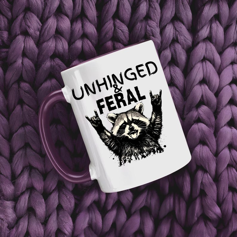 Puede incluir: Taza de cer&aacute;mica blanca con asa morada. Presenta una ilustraci&oacute;n en blanco y negro de un mapache haciendo un gesto de rock and roll con la mano, con el texto "UNHINGED & FERAL" encima.