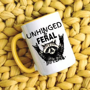 Puede incluir: Taza de cer&aacute;mica blanca con asa y borde amarillos. La taza presenta el texto "UNHINGED & FERAL" sobre una ilustraci&oacute;n en blanco y negro de un mapache haciendo un gesto de rock and roll.