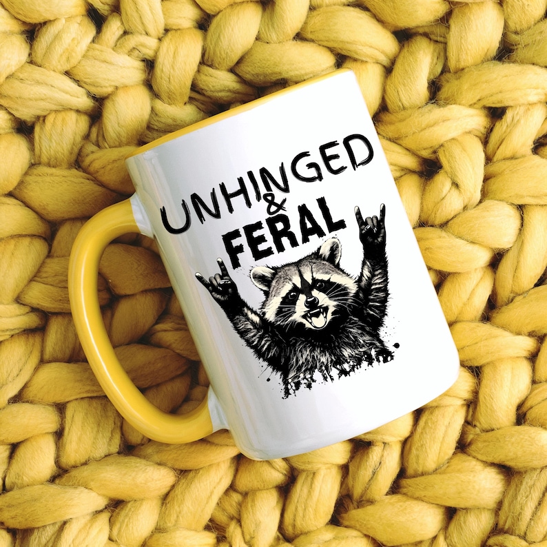 Puede incluir: Taza de cer&aacute;mica blanca con asa y borde amarillos. La taza presenta el texto "UNHINGED & FERAL" sobre una ilustraci&oacute;n en blanco y negro de un mapache haciendo un gesto de rock and roll.