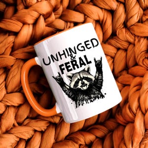 Puede incluir: Taza de cer&aacute;mica blanca con asa y borde naranja. La taza presenta el texto "UNHINGED & FERAL" sobre una ilustraci&oacute;n en blanco y negro de un mapache haciendo un gesto de rock and roll.