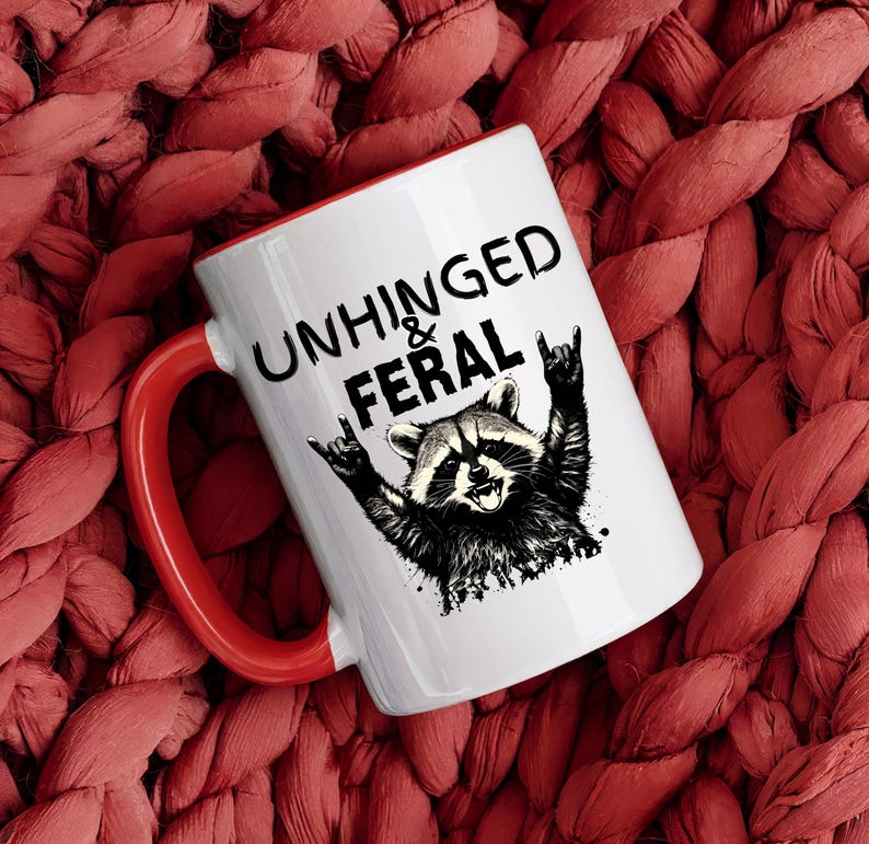 Puede incluir: Taza de cer&aacute;mica blanca con interior y asa rojos. La taza presenta el texto "UNHINGED & FERAL" sobre una ilustraci&oacute;n en blanco y negro de un mapache haciendo un gesto de rock and roll.