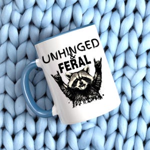 Puede incluir: Taza de cer&aacute;mica blanca con asa y borde azules. La taza presenta una ilustraci&oacute;n en blanco y negro de un mapache con los brazos levantados en un gesto de rock and roll. Las palabras "UNHINGED & FERAL" est&aacute;n impresas encima del mapache.
