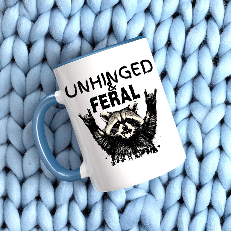 Puede incluir: Taza de cer&aacute;mica blanca con asa y borde azules. La taza presenta una ilustraci&oacute;n en blanco y negro de un mapache con los brazos levantados en un gesto de rock and roll. Las palabras "UNHINGED & FERAL" est&aacute;n impresas encima del mapache.
