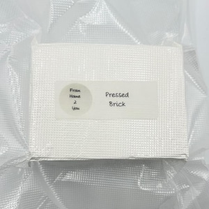 May include: A white, rectangular pressed brick, packaged in clear plastic. The brick has a textured surface. A white label on the brick reads "Pressed Brick" and "From Home 2 You".