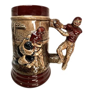 May include: A ceramic beer stein with a football theme. The stein is brown and burgundy with relief images of football players. The handle is a sculpted football player in a maroon uniform catching a football. The stein has a wide base.