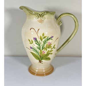 May include: Cream-colored ceramic pitcher with a green handle and rim. The pitcher features a floral design with green leaves, purple and pink flowers. The base is a light brown color. A decorative piece for the kitchen or dining room.
