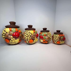 Puede incluir: Juego de cuatro botes de cerámica con tapas. Los botes están decorados con un diseño de frutas texturizado en tonos amarillos, naranjas, marrones y rojos. Las tapas son de color marrón oscuro.