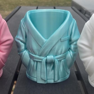 May include: Three ceramic robe-shaped planters in pink, teal, and white. Each planter features a robe design with a belt and pockets. The planters are arranged side by side on a dark surface, showcasing their unique shapes and colors.